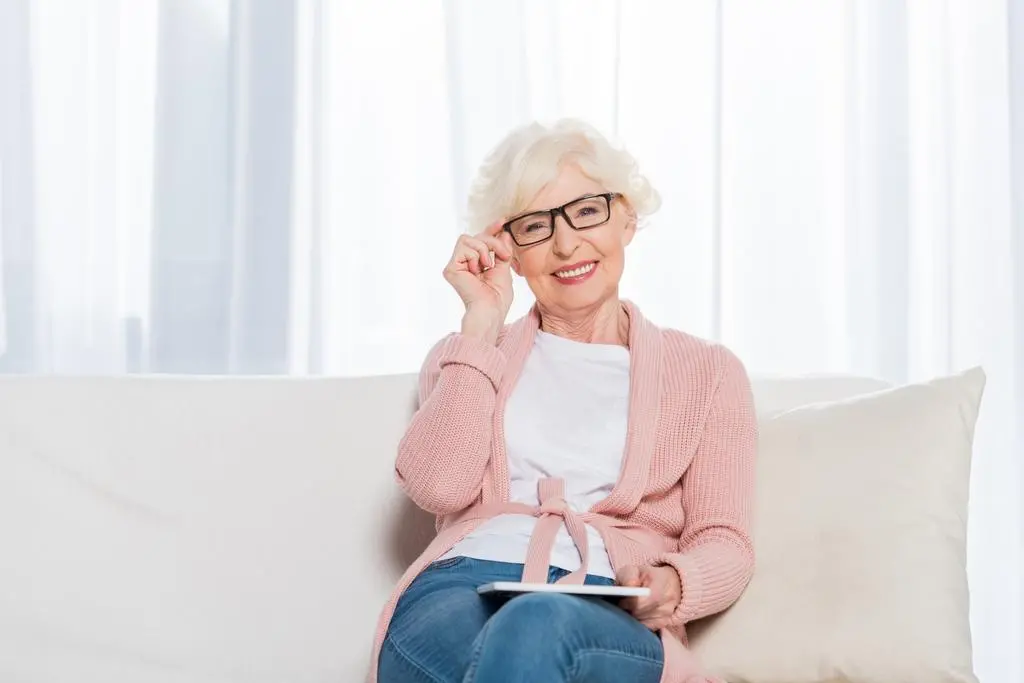 Idősebb nő mosolyogva igazítja a szemüvegét a kanapén ülve, kezében tablet.