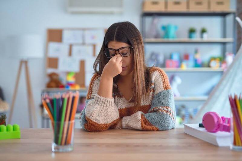 Fiatal nő pulóverben ül az iskolában, orrnyergét dörzsöli a szemüvegét feltolva